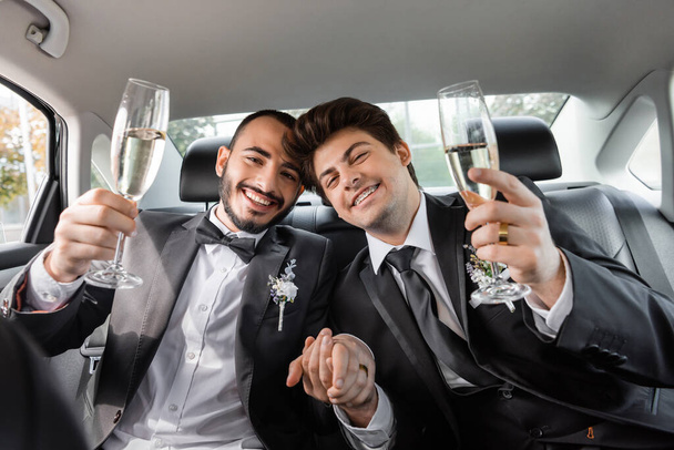 Smiling young gay newlyweds in formal wear holding hands and champagne while looking at camera during road trip while sitting on backseat of car  - Photo, Image