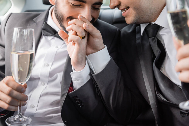 Smiling and bearded gay man with wedding ring on hand kissing hand of boyfriend in braces and holding blurred champagne while sitting in car during honeymoon  - Photo, Image