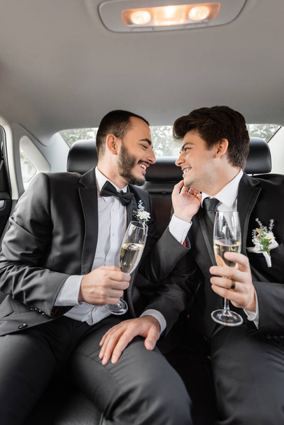 Side view of smiling and bearded gay groom in classic suit touching young boyfriend in braces with glass of champagne while celebrating wedding on backseat of car during honeymoon  - Zdjęcie, obraz