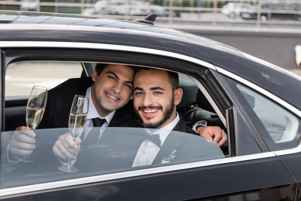 Uśmiechnięte panny młode tej samej płci w formalnym ubraniu trzymające kieliszki szampana i patrzące w kamerę przez okno siedząc na tylnym siedzeniu samochodu przed wyjazdem na miesiąc miodowy  - Zdjęcie, obraz