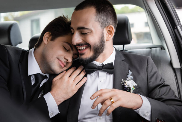 Homem barbudo sorridente em terno elegante com boutonniere sentado perto do jovem noivo morena com olhos fechados no carro após a celebração do casamento enquanto vai em lua de mel  - Foto, Imagem