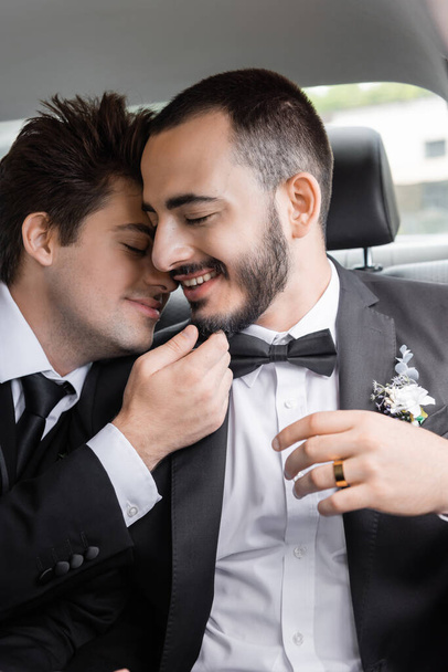 Mladá brunetka gayové ženich v elegantní formální oblečení dotýkat bradu vousatý přítel se zavřenýma očima po svatební oslavě při odchodu na líbánky  - Fotografie, Obrázek