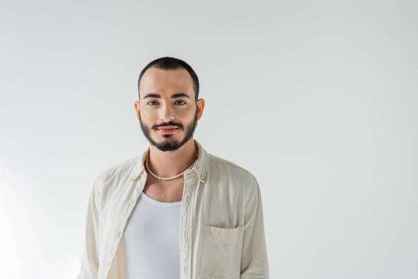 Portrait of young short haired and bearded homosexual man in pearl necklace and casual clothes looking at camera while standing isolated on grey   - Photo, Image