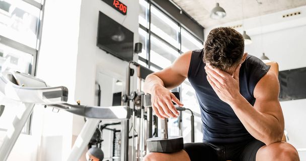Exhausted muscular male athlete in sportswear sitting with dumbbell and touching forehead while resting after workout in modern gym - Photo, Image