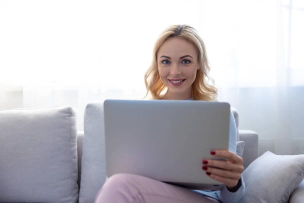 Pleased pretty blonde lady seated on the cozy sofa with her laptop looking before her - Φωτογραφία, εικόνα