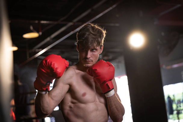 νεαρός άνδρας με μυώδες σώμα γάντια πυγμαχίας σε ένα γυμναστήριο και κάμψη των μυών - Φωτογραφία, εικόνα