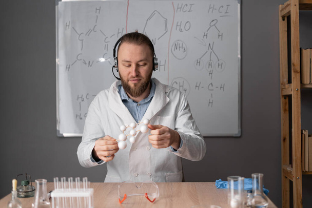 Ein bärtiger Chemielehrer mit weißem Headset sitzt auf einem Stuhl vor dem Hintergrund einer Markierungstafel mit Formeln und hält eine Molekülprobe in seinen Händen. Online-Tutorenkonzept. Webcam ansehen. - Foto, Bild