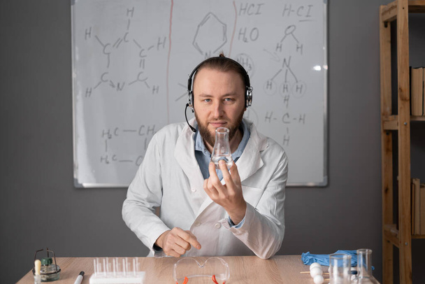 ένας γενειοφόρος καθηγητής χημείας με λευκά ακουστικά κάθεται σε καρέκλα στο φόντο πινακίδας με τύπους, κρατώντας μια φιάλη υγρού στα χέρια του. online διδασκαλία έννοια. Προβολή κάμερας. - Φωτογραφία, εικόνα