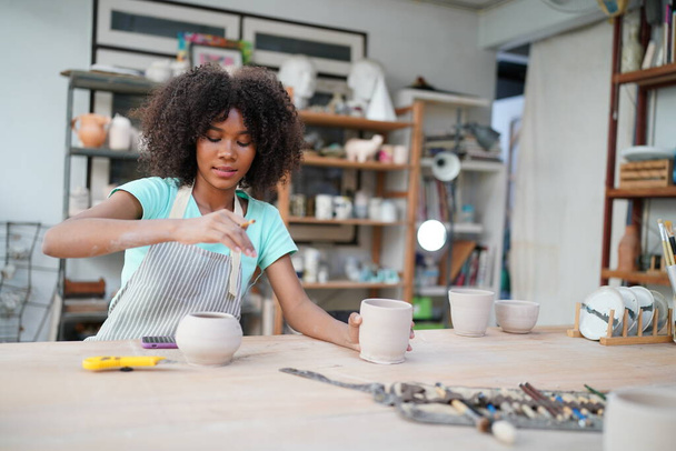 Młoda Afro dziewczyna garncarz ręczny podejmowania gliny wazon w warsztacie garncarskim, Właściciel firmy.  - Zdjęcie, obraz