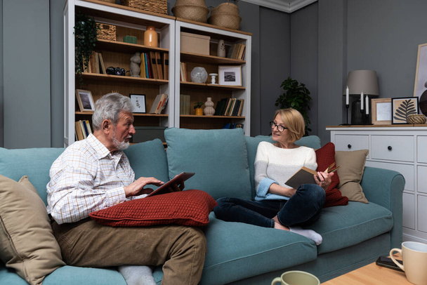 Happy senior married couple relaxing together at home with the wife sitting back from her husband as she reads a book while he surfs the internet on a tablet. Love and respect in older age concept - Zdjęcie, obraz