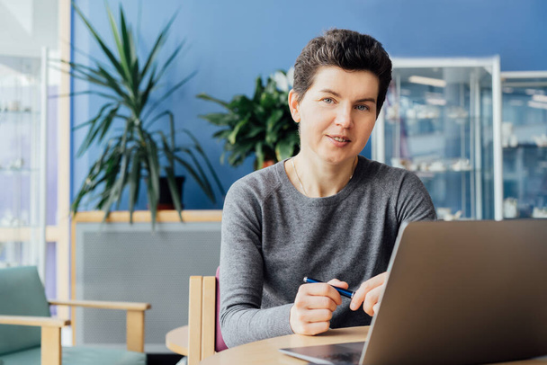 Neutral gender middle aged woman with no make-up in casual clothing using laptop and smiling while working indoors in her workstation in an open space office. Online video call, IT HR, recruiting. - Фото, зображення