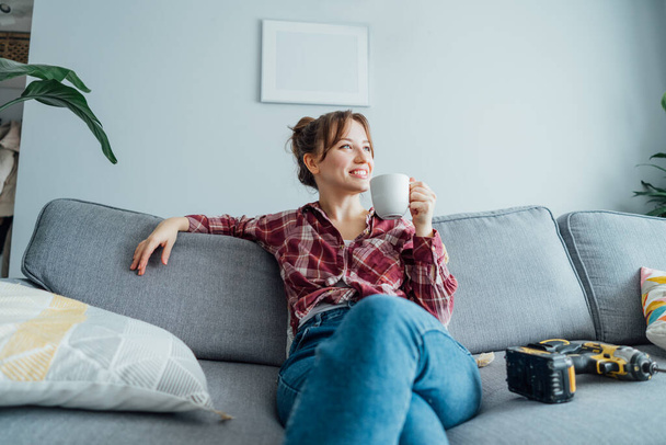 Happy smiling proud of herself young woman relaxing with coffee cup after she puts by her own a picture on the wall at her home. Housekeeping work. Doing repair herself. DIY, gender equality concept. - Φωτογραφία, εικόνα