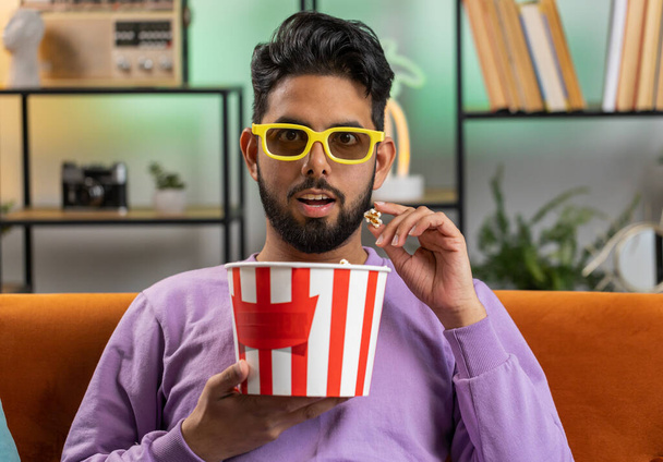 Excited indian man in 3D glasses sits on sofa eating popcorn snacks and watching interesting TV serial sport game, film, online social media movie content at home apartment. Guy enjoying entertainment - Photo, Image