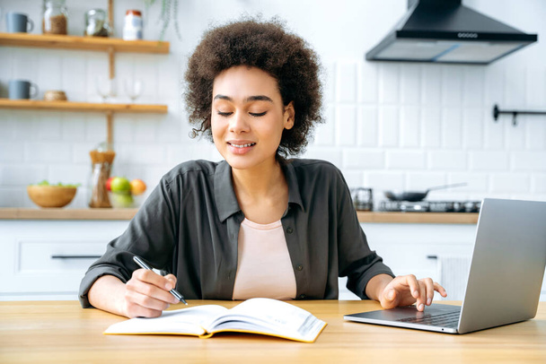 Remote work or education. Positive african american girl with short curly hair, sit at a table in a kitchen, using a laptop, taking notes in notebook, working remotely, studying, doing homework, smile - Foto, Imagem