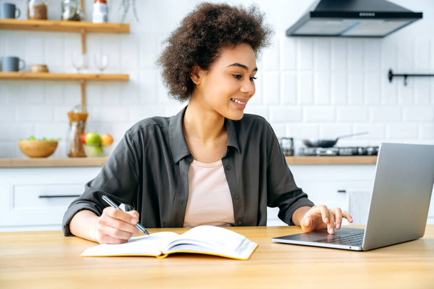 Positive stylish modern african american girl with short curly hair, sitting at a table in the kitchen, using a laptop, taking notes in a notebook, working remotely, studying, doing homework, smile - Fotó, kép