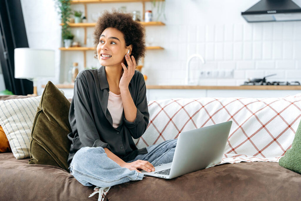 Happy pretty, curly haired african american woman, in stylish wear, sit on sofa in living room with laptop, spend leisure time on the Internet, listening online webinar or music using earphone, smile - Photo, Image