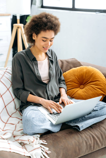 Foto vertical de la joven afro-americana de pelo rizado, estudiante, freelancer, programador, se sienta en un sofá en la sala de estar con un ordenador portátil, trabajando en línea, escribiendo en un teclado, sonrisas - Foto, imagen