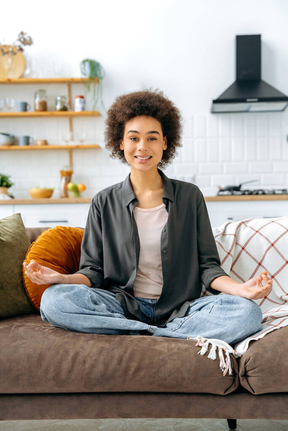 Vertical photo of calm happy peaceful relaxed african american young woman, sits alone at home in living room on sofa and meditating in the lotus position, smile at camera. Relaxation and meditation - Fotó, kép