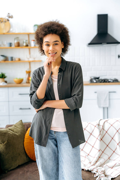 Vertical photo of a gorgeous positive stylish african american girl with short curly hair, stand in living room at home against the background of the kitchen, looking at the camera, smiles friendly - Photo, Image