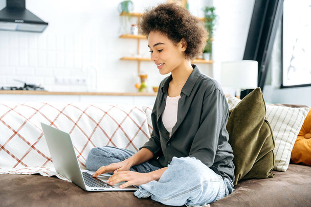 Fernarbeit oder Studium. Positiv wunderschöne junge afrikanisch-amerikanische lockige Frau, Studentin oder Freiberuflerin, sitzt auf einem Sofa im Wohnzimmer, nutzt Laptop für Online-Arbeit oder Lernen, lächelt freundlich - Foto, Bild