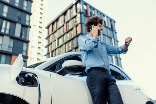 Progressive businessman talking on the phone, leaning on electric car recharging with public EV charging station, apartment condo residential building on the background as green city lifestyle. - Photo, Image