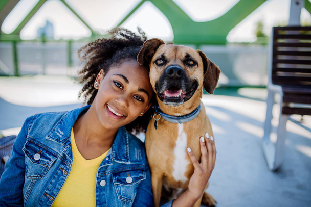 Portrait of happy multiracial girl with her dog, sitting outdoor. Concept of relationship between dog and teenager, everyday life with pet. - Фото, изображение
