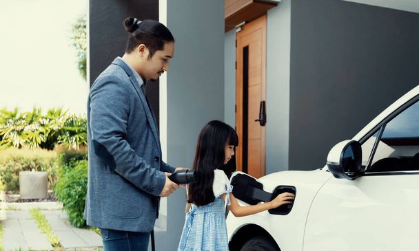 Progressive father and daughter plugs EV charger from home charging station to electric vehicle. Future eco-friendly car with EV cars powered by renewable source of clean energy. - Foto, immagini
