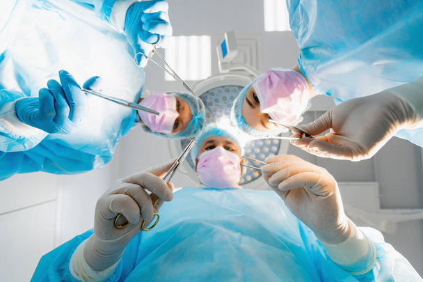 Ομάδα ιατρών χειρουργών και νοσηλευτών που κατέχουν νυστέρια χειρουργικά εργαλεία κατά τη διάρκεια της χειρουργικής επέμβασης στο κάτω μέρος του νοσοκομείου - Φωτογραφία, εικόνα
