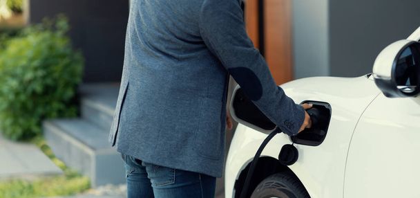 Closeup progressive asian man install cable plug to his electric car with home charging station. Concept of the use of electric vehicles in a progressive lifestyle contributes to clean environment. - Φωτογραφία, εικόνα