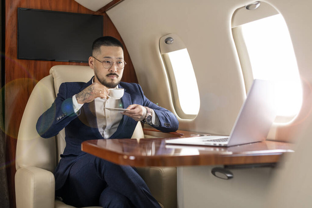 Başarılı Asyalı iş adamı özel uçakta oturup kahve içiyor ve dizüstü bilgisayara bakıyor. Gözlüklü Koreli finansör ve iş kıyafetleri içinde uçuyor. Lüks bir yaşam tarzı. - Fotoğraf, Görsel