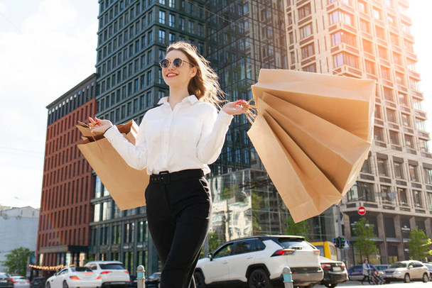 attraktives junges Mädchen in Sonnenbrille und weißem Hemd hält Papiertüten mit Einkäufen auf der Straße in der Stadt, Frau geht mit Taschen einkaufen und lächelt, Einkaufskonzept - Foto, Bild
