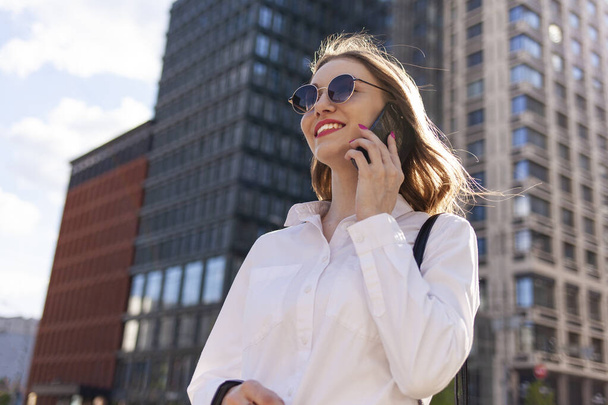 young attractive girl in sunglasses and white shirt is talking on the phone and smiling on the street in the city, woman is talking on mobile phone and walking outdoors - Photo, image