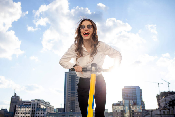 young happy girl in sunglasses rides forward on electric scooter and shouts in the street, woman uses eco transport and rejoices in the city - Фото, изображение