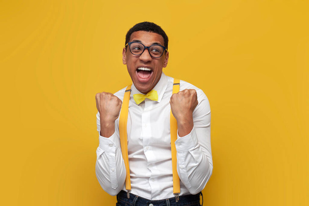 young African American guy in white shirt with suspenders and bow tie rejoices in victory on yellow isolated background, nerd man in glasses wins and dances - Фото, изображение