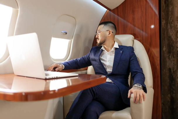 успешный азиатский бизнесмен в костюме и очках сидит в частном самолете с ноутбуком и смотрит в окно, корейский предприниматель в деловой одежде летает в самолете, роскошный образ жизни - Фото, изображение