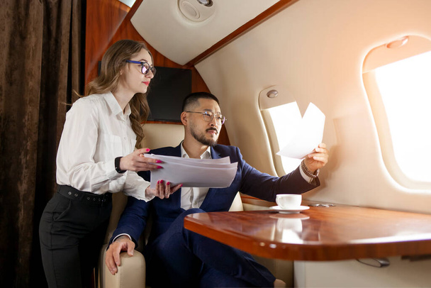 aziatische zakenman met vrouwelijke collega in pak en bril zit in prive-jet en kijkt naar documenten, Koreaanse ondernemer vliegt in het vliegtuig en werkt met papieren, luxe levensstijl - Foto, afbeelding