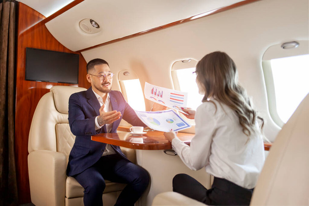 obchodní jednání v letadle, asijský podnikatel v soukromém tryskáči mluví s kolegyní a prohlíží si grafy a dokumenty, podnikatelé létají v letadle a pracují s papíry - Fotografie, Obrázek