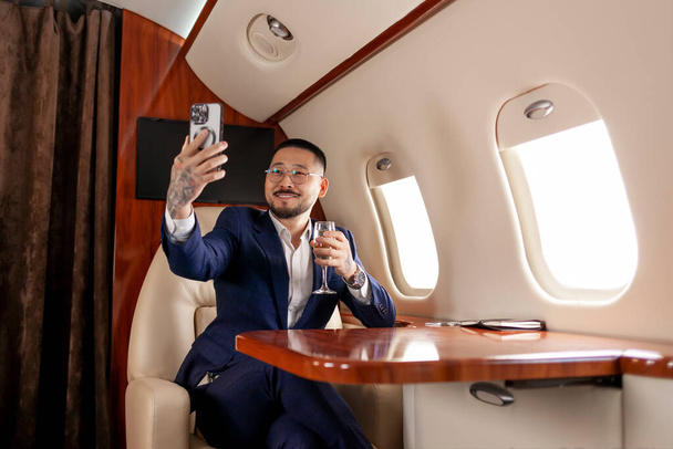 επιτυχημένος Ασιάτης επιχειρηματίας με κοστούμι και γυαλιά κάθεται σε ιδιωτικό τζετ με ένα ποτήρι σαμπάνια και επικοινωνεί μέσω βιντεοκλήσης, Κορεάτης επιχειρηματίας πετάει με αεροπλάνο και βγάζει selfie, luxury lifestyle - Φωτογραφία, εικόνα