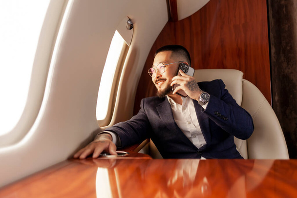 азиатский бизнесмен в костюме сидит в частном самолете и разговаривает по телефону, корейский предприниматель летает на самолете и общается по мобильному телефону - Фото, изображение