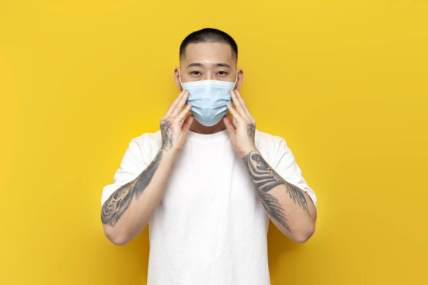 Asian man in medical mask and white football stands on yellow isolated background, Korean guy in protective mask against viruses and flu - Photo, Image