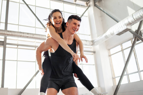 young happy athletic couple in sportswear runs and rejoices in bright gym, the guy carries the girl on his back and smiles, the concept of freedom - Фото, изображение