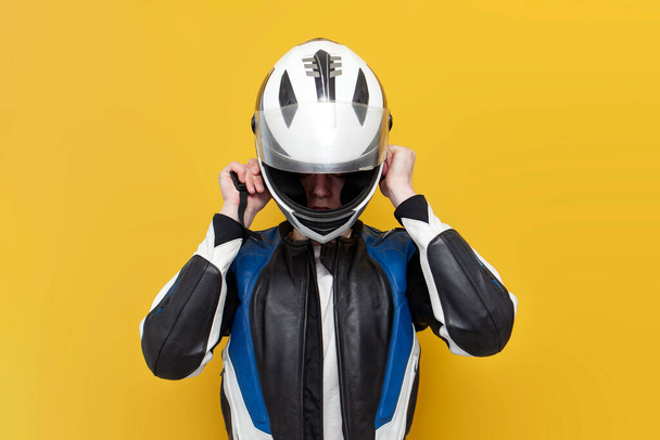 young guy motorcyclist in leather jacket puts on helmet on yellow isolated background, professional racer in protective gear holds helmet, copy space - 写真・画像