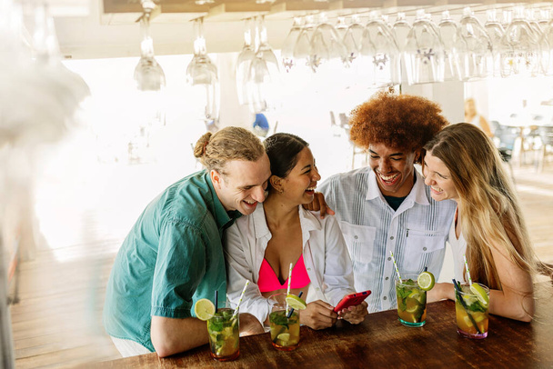 Jonge groep van gelukkige vrienden die plezier hebben met behulp van mobiele telefoon terwijl ze genieten van mojito cocktail drankje aan het strand bar - Foto, afbeelding