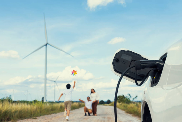 Concept of progressive happy family holding windmill toy and relax at wind farm with electric vehicle. Electric vehicle driven by clean renewable energy from wind turbine generator to charger station. - Foto, Imagem
