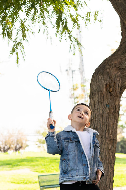 Aktywne dziecko bawiące się w badmintona w Sunny City Park, bawiące się rakietą i wahadłowcem na Green Grass Field Against Blue Sky. Kid uderzając Shuttlecock z rakiet. - Zdjęcie, obraz