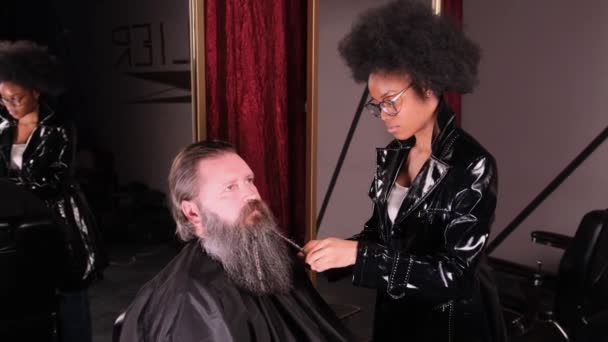 Una mujer afroamericana con estilo en un impermeable negro en una peluquería trenzas sus trenzas en una barba para un cliente masculino. - Imágenes, Vídeo