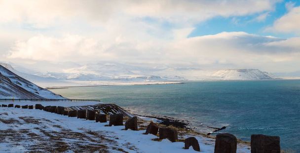 A panoramic shot of stones barriers before a blue sea surrounded by hills covered in snow - Фото, зображення