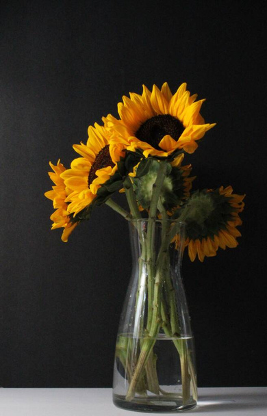 A vertical shot of a bunch of sunflowers in a glass vase on the table in the dark background - Zdjęcie, obraz