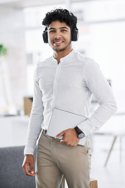 Dressing professionally doesnt have to include a suit. Portrait of a young businessman wearing headphones and holding a laptop in an office - Foto, Bild