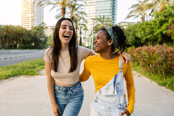 Two multiracial female friends laughing together outdoors. Young women embracing each other having fun together walking in city street. - Фото, изображение